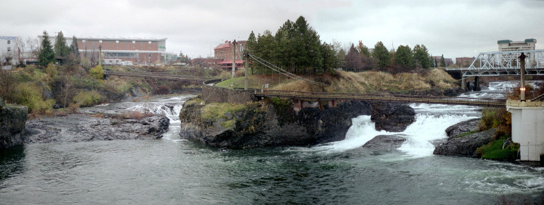 [The bridge from the prior photo is in the background on the right. Most of the water flow and froth is on the right side of the island in the middle of the river. The island is quite a ways above the water line and has buildings on it.]